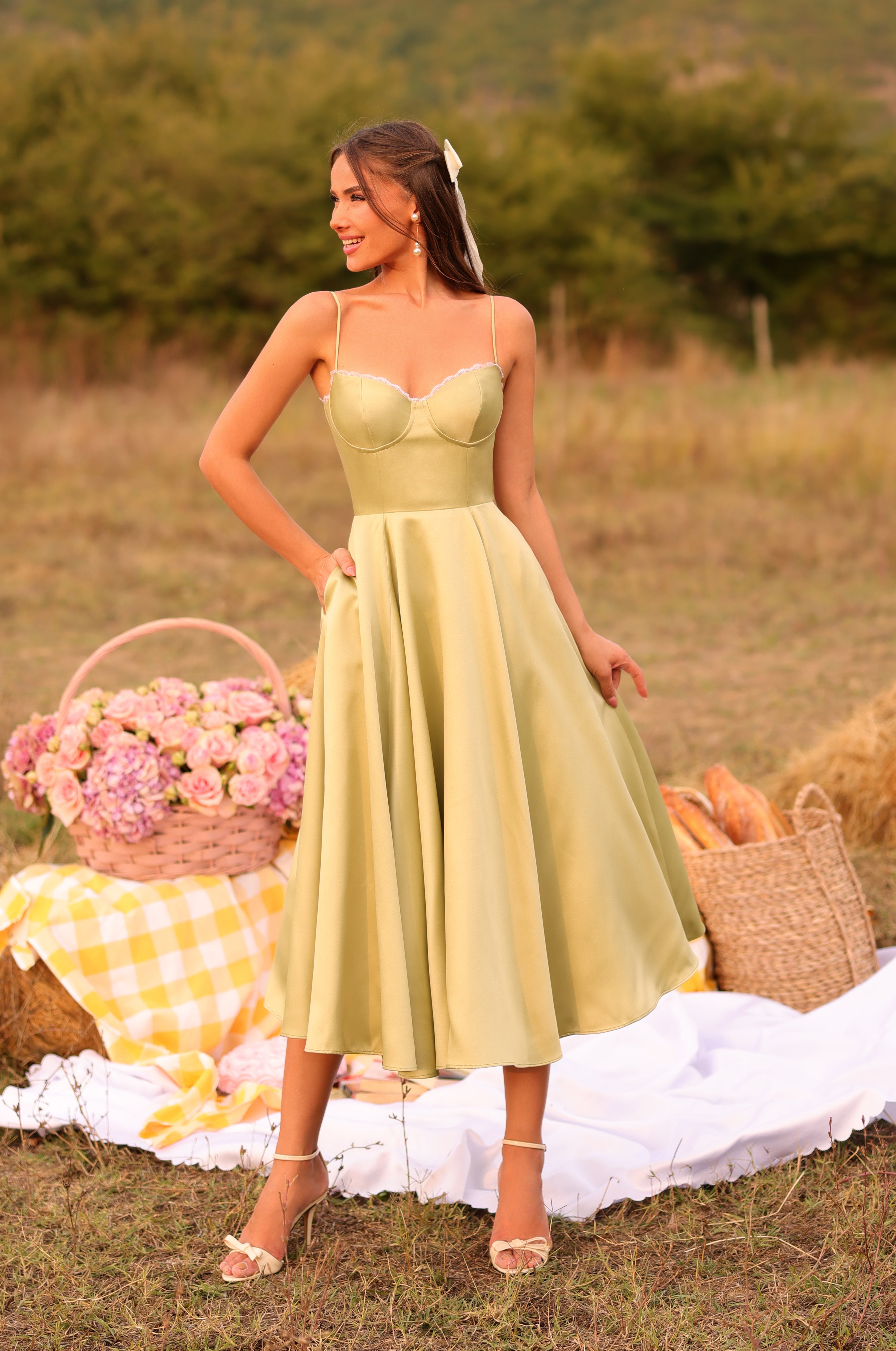 A woman in the Olivetta Olive Green Satin Midi Wedding Guest Dress stands outdoors on a picnic setup with flowers, bread, and a yellow-checkered cloth on the grass.